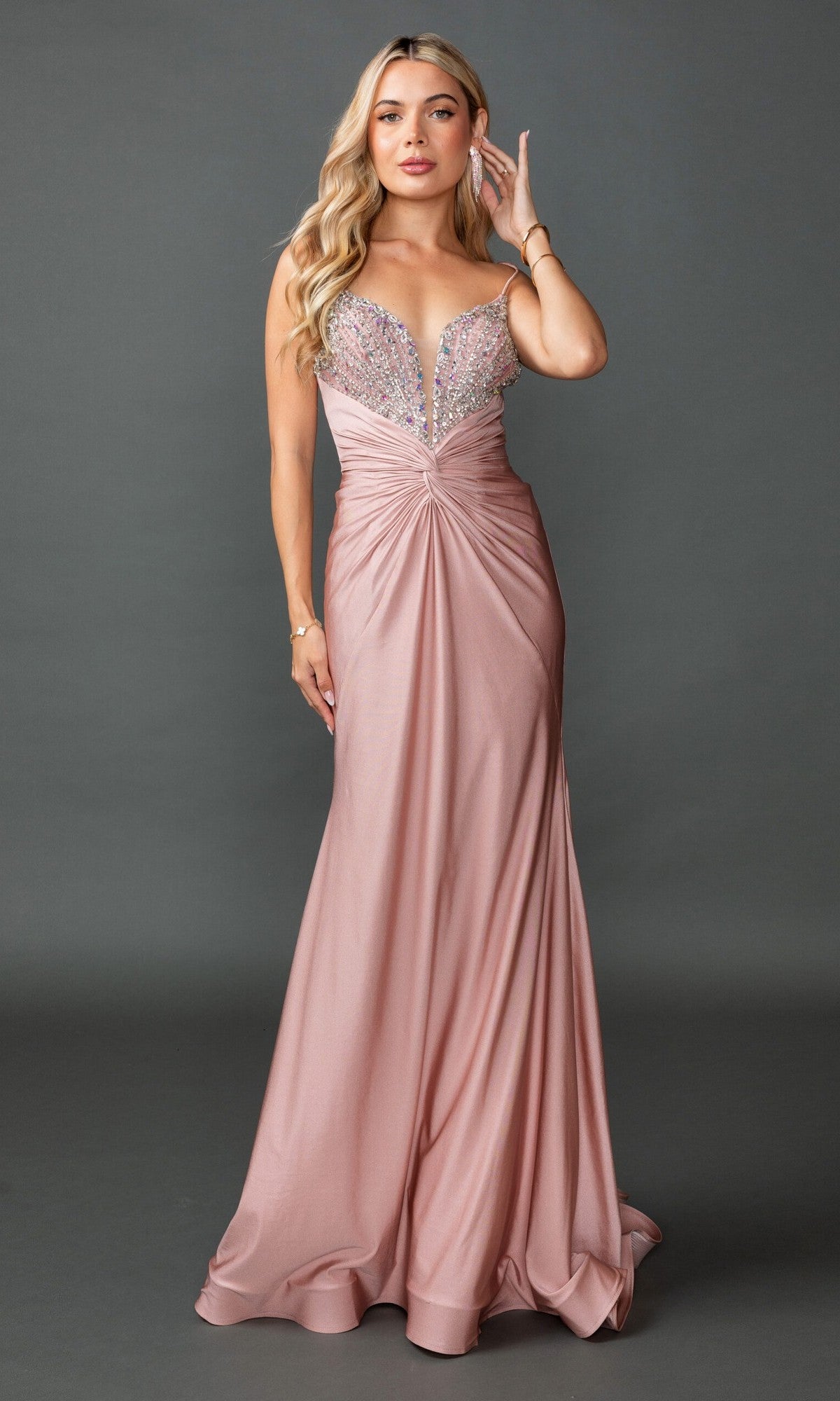 A woman poses against a gray background wearing the Dancing Queen 4553, a lace-up long pink prom dress featuring a twist-front, sparkly deep V-neck bodice, thin straps, and beaded open back.